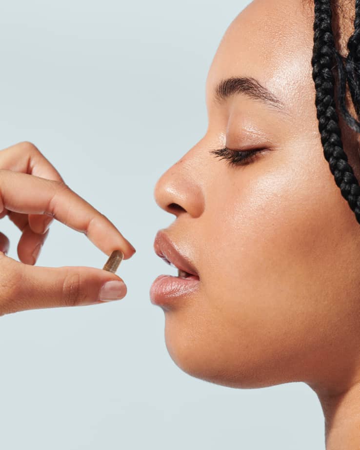 Woman photopraphed from the side holding a supplement pill about to put it in her mouth on light blue beige background