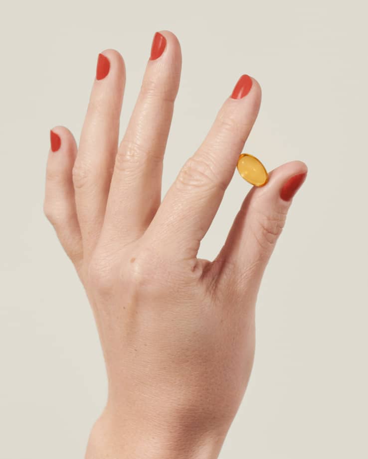 Hand with red nail polish holding a supplement pill in the air on a beige background