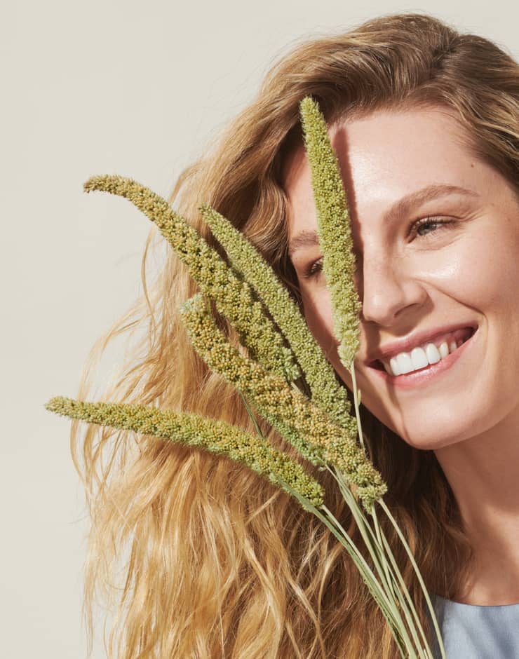 Blong wavy haired woman similing holding a few pieces of millet in front of her face on beige background