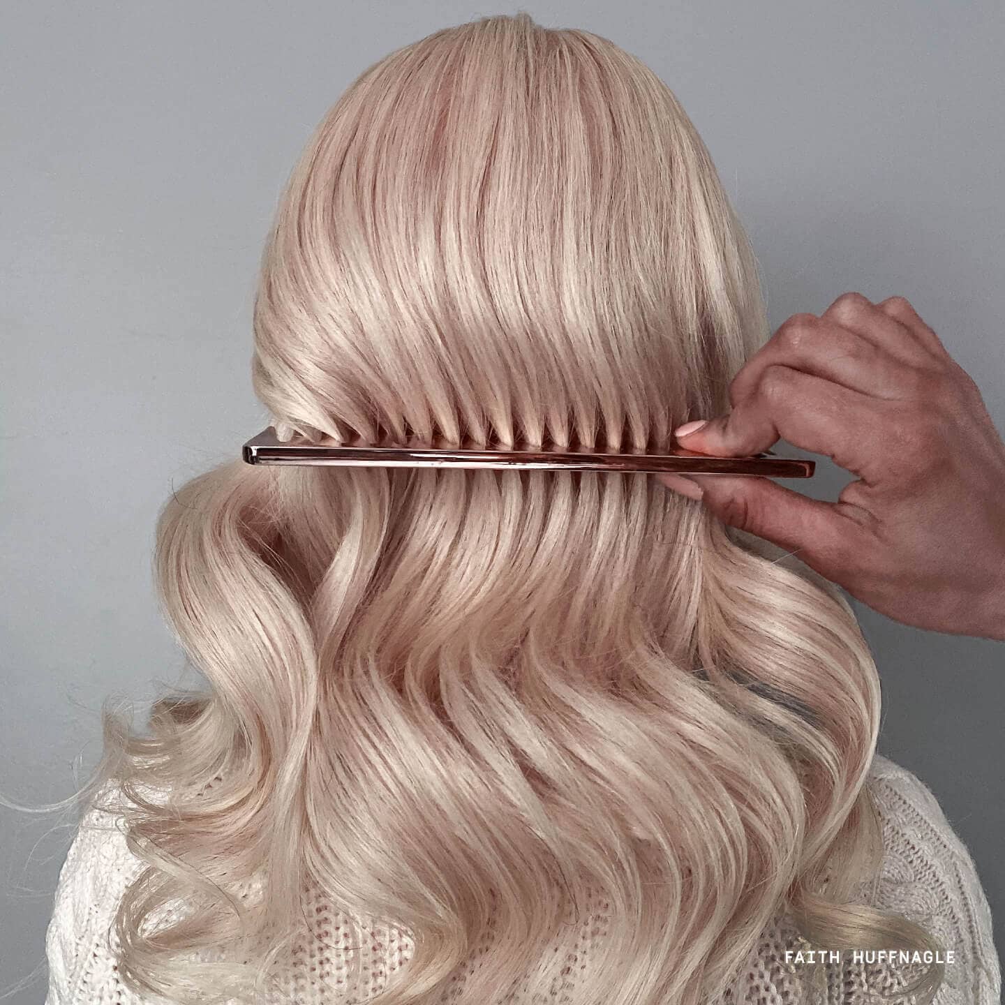 Blonde hair woman with gold comb raking through hair in front of grey background